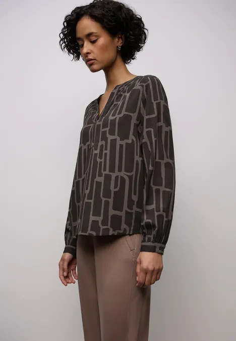 Dame trägt graue Bluse mit abstraktem Rechteckmuster und beige Hose.